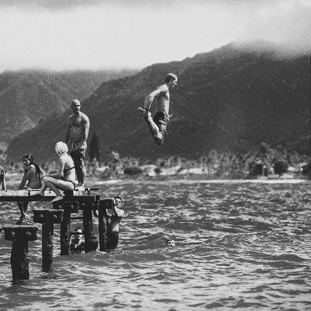 Black & white image of: Man, probably in his late teens or early twenties, jumping off a dock into a lake. 