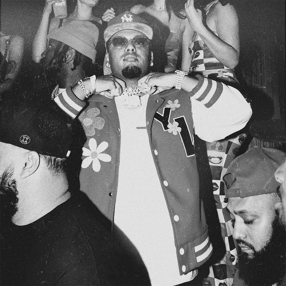 Black & white image of: A a guy in a baseball cap and tinted glasses holding the gold chain around his neck at what looks to be a hip hip party / event. 